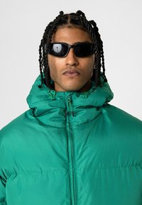 Groene gewatteerde jack met een hoge kraag, voorzien van een ritsluiting en verstelbare touwtjes. Gestyled met zwarte zonnebril en een soepele textuur.