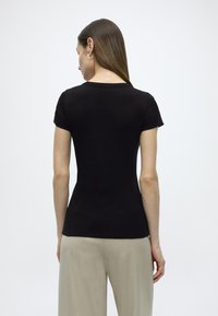 Μαύρο εφαρμοστό T-shirt με κοντά μανίκια και στρογγυλή λαιμόκοψη. Μαλακή, λεία υφή, η πίσω όψη δείχνει ελάχιστες ραφές. Συνδυασμένο με ανοιχτόχρωμο παντελόνι.