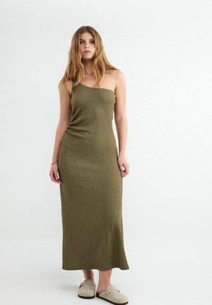Kobieta w teksturowanej oliwkowo-zielonej sukience na jedno ramię bez rękawów i beżowych butach wsuwanych, stoi na tle jednolitego jasnego tła.