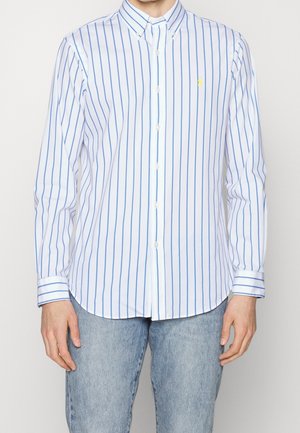 Homme portant une chemise blanche à rayures verticales bleues et un jean bleu clair, debout devant un fond uni.