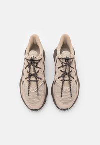 Pár béžových a hnědých sneakers vyrobených z vícebarevné síťoviny s tmavě hnědými tkaničkami upevněnými černými zámky na tkaničky, zobrazené z vrchu na bílém pozadí.