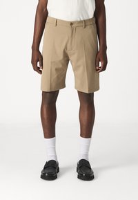 Beige maßgeschneiderte Shorts mit einem frontalen Bundfalten, zwei Seitentaschen und einem Knopfverschluss. Getragen mit weißen Socken und schwarzen Slippern.