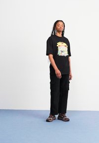 Svart grafisk t-skjorte med fargerik print, sammen med svarte cargo-bukser og brune sneakers, stående mot en lys bakgrunn.