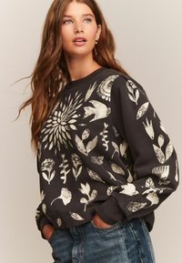 Kvinde med langt brunt hår iført sort sweatshirt med hvide blomster- og fuglemønstre, hænderne i lommerne, ser let opad.