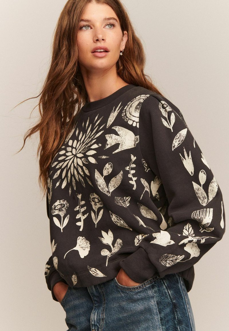 Kvinde med langt brunt hår iført sort sweatshirt med hvide blomster- og fuglemønstre, hænderne i lommerne, ser let opad.