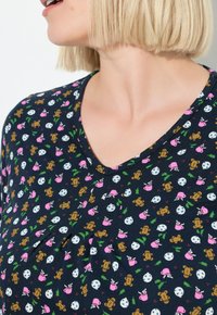 Frau mit kurzem blonden Haar, die ein navyblaues V-Ausschnitt-Oberteil trägt, das mit Lebkuchenmännern, rosa Tassen, Schneeflocken und Stechpalmenblättern gemustert ist.