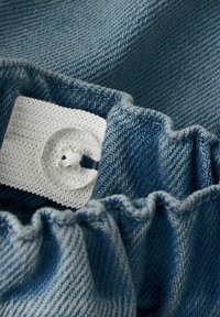 Krupan plan plavog trapera s elastičnim pojasom i bijelim podesivim gumbom na prišivenom tkaninskom trakici.