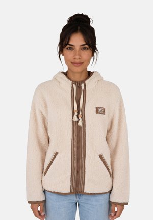 Mujer con cabello oscuro recogido lleva una chaqueta sherpa beige con cremallera, ribetes marrones, cordones y bolsillos delanteros sobre vaqueros azul claro.