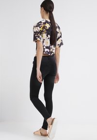 Černé skinny džíny v kombinaci s květinovým topem, který má krátké rukávy, na bílém pozadí. Sandály s béžovým páskem.