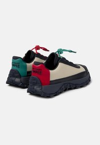 Zapatillas deportivas con parte superior de lienzo beige, detalles en negro, detalles en el talón en rojo y verde, y una suela negra gruesa con un patrón de agarre texturizado.