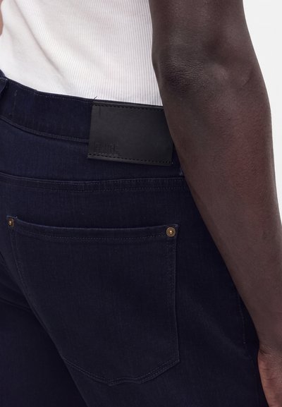 Σκούρα μπλε τζιν από denim με πίσω τσέπη και μαύρη ετικέτα, φορεμένα με λευκό ριμπ μπλουζάκι χωρίς μανίκια, που δείχνει μέρος του χεριού και της μέσης του φορέα.