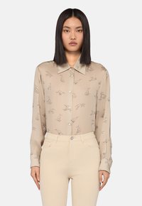Donna che indossa una camicia beige con bottoni e illustrazioni di piccoli cani, insieme a pantaloni color crema, in piedi contro uno sfondo chiaro uniforme.