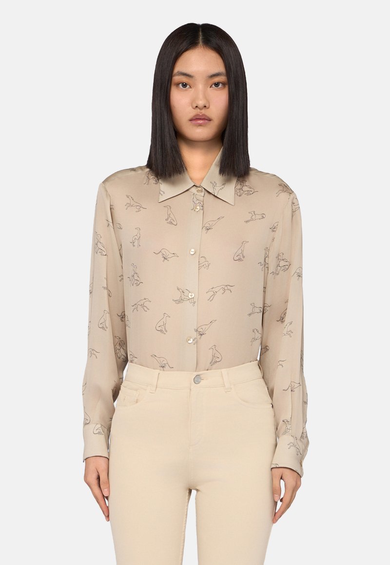Donna che indossa una camicia beige con bottoni e illustrazioni di piccoli cani, insieme a pantaloni color crema, in piedi contro uno sfondo chiaro uniforme.