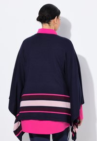 Frau mit dunklem Haar im niedrigen Dutt, trägt große Hoop-Ohrringe, eine marineblaue Capa mit rosa und lila Streifen sowie ein hellrosa Shirt.