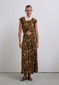 Vestido maxi de estampado marrón y crema con mangas cortas, cintura ajustada que presenta dos cortes circulares, combinado con una bufanda blanca y sandalias.