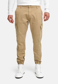 Homme portant un pantalon cargo beige avec des poignets élastiques et des baskets montantes blanches, sur un fond uni.