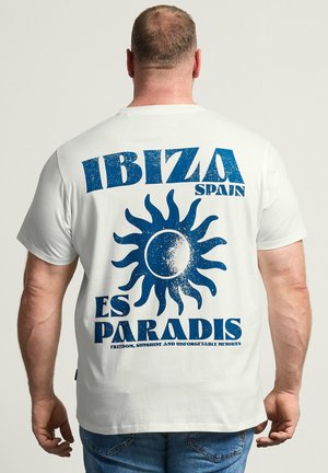 Mann trägt weißes T-Shirt mit blauem Sonnenmotiv und dem Text „IBIZA SPAIN ES PARADIS“ auf dem Rücken, steht vor einfarbigem Hintergrund.