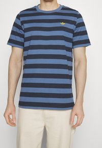 Man i en blå och svart horisontellt randig t-shirt med kort ärm och en liten gul logotyp på bröstet, i kombination med beiga byxor.