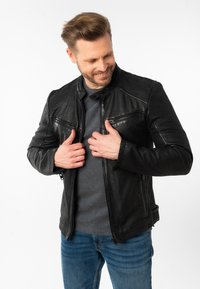 Giacca di pelle nera con zip frontale, spalle rigate e due tasche sul petto, indossata sopra una camicia grigia e jeans blu. Tessuto liscio.