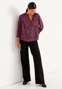 Paarse kanttop met een V-hals en 3/4 mouwen, gecombineerd met zwarte wijde broek en zwarte open-teen schoenen. De textuur heeft bloemmotieven.
