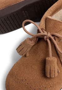 Zapato de ante marrón con puntera redondeada, costuras decorativas y acentos de borlas en los cordones. Suela de goma con un patrón texturizado.