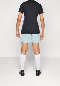Zwart sportshirt met kleurrijke zijaccenten, lichtblauwe korte broek, witte sokken en zwarte voetbalschoenen met gouden details.