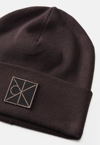 Gorro de malha castanho com punho dobrado, apresentando um patch retangular preto com detalhes de logotipo bordados. Textura suave e design ajustado.
