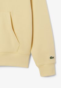 Sudadera amarilla con un bolsillo tipo canguro, puños y dobladillo acanalados. Presenta un pequeño logotipo de cocodrilo verde en la manga izquierda. Textura suave.
