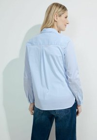 Light blue striped shirt with a collar, long sleeves, and a back pleat detail. Fabric appears smooth; shirt is paired with dark denim jeans.