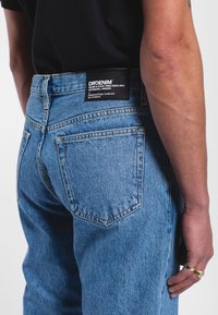 Modré džínové kalhoty s vysokým pasem, které mají černou značkovou nášivku blízko zadního pásku a dvě zadní kapsy. Tkanina má texturovaný povrch.