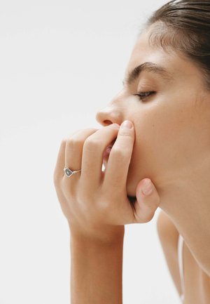 Anillo de plata con un diseño geométrico que presenta piedras preciosas oscuras, llevado en una mano que toca la cara, mostrando piel suave y tonos naturales.