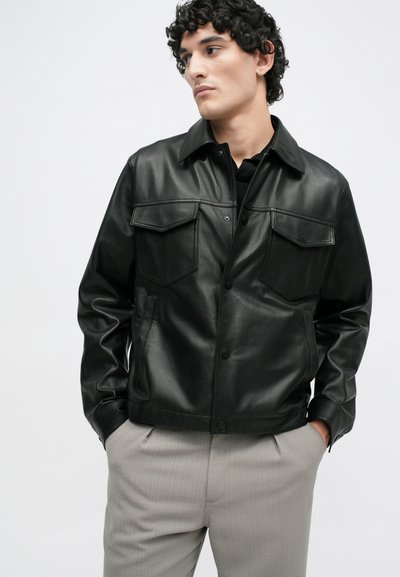 Μαύρο δερμάτινο μπουφάν με αιχμηρό γιακά, δύο τσέπες στο στήθος και κλείσιμο με κουμπί. Φορεμένο πάνω από ένα μαύρο πουκάμισο και γκρι παντελόνι. Λείος υφή.