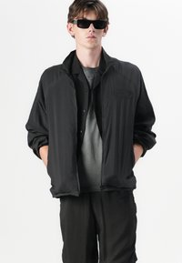 Veste zippée noire légère avec un col haut, présentant une finition texturée et un logo brodé sur la poitrine, assortie de shorts sombres.