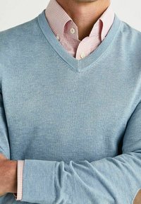 Jasnoniebieski sweter z dekoltem w serek nałożony na różowo-białą koszulę w kratę gingham; połączenie tekstur i kolorów tworzy elegancki, casualowy look.