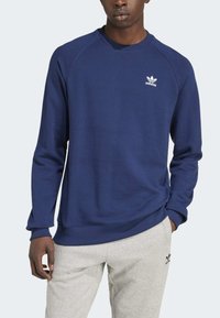 Navy zila krekla ar garām piedurknēm, kas izgatavots no kokvilnas, ar ribotām apkaklītēm. Uz krūtīm ir neliels balts Adidas logo.