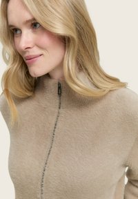 Beige ritsweater met een hoge kraag, gemaakt van zachte, gestructureerde stof. Heeft een subtiele naad die langs de voorkant loopt en een ritstrekkertje.