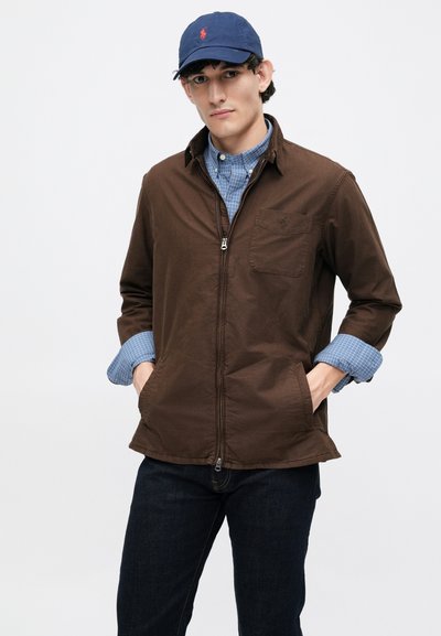 Casaco castanho com fecho éclair, com um bolso no peito, combinado com uma camisa xadrez azul clara e jeans de ganga escura. Boné azul-marinho com logótipo vermelho.