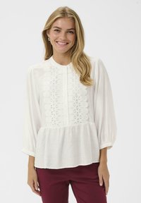 White blouse with long balloon sleeves, intricate lace detailing down the front, and a gathered bottom hem. Paired with maroon pants.