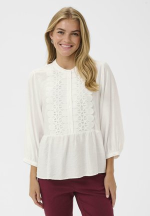 White blouse with long balloon sleeves, intricate lace detailing down the front, and a gathered bottom hem. Paired with maroon pants.