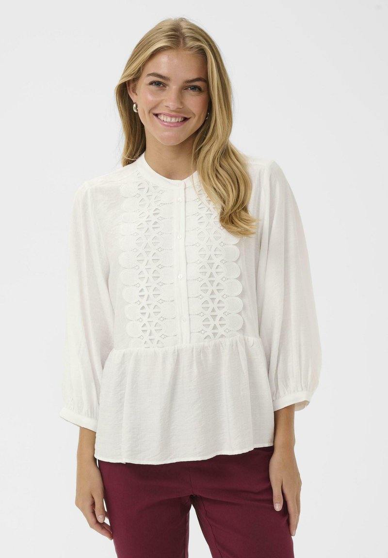 White blouse with long balloon sleeves, intricate lace detailing down the front, and a gathered bottom hem. Paired with maroon pants.