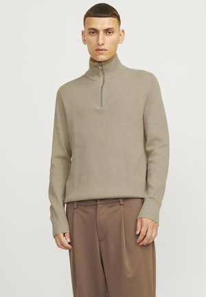 Beige bordázott kötött pulóver magas gallérral és fél zipzárral, barna plissé nadrággal párosítva. Simaság és testhezálló fazon jellemzi.