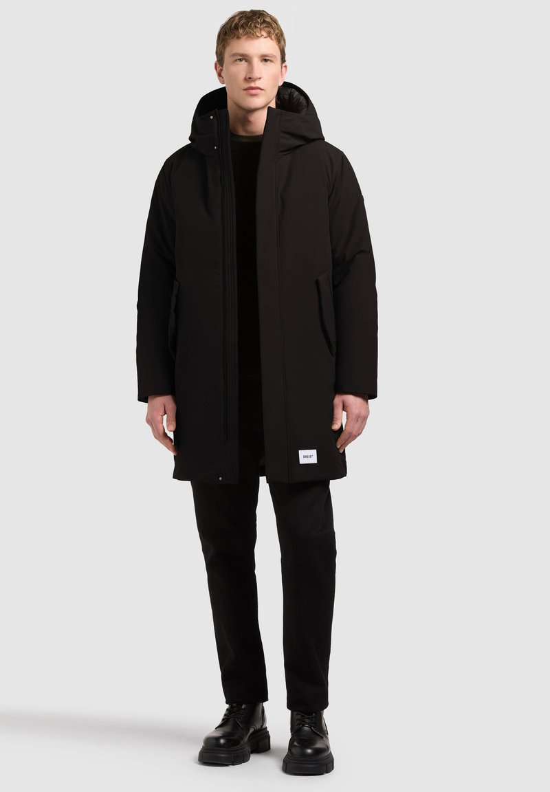 Homme portant un long manteau d'hiver noir à capuche avec poches latérales, pantalon noir et bottes noires à lacets, debout devant un fond uni.
