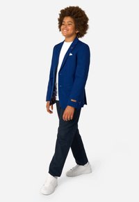 Zila žakete ar vienu krūšu kabatu, ar zeltītu "DAILY" tekstu uz piedurknes, apvienota ar balto kreklu un tumšiem biksēm, valkāta ar baltām kedām.