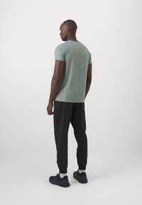 Grünes Heide-T-Shirt, kurzärmelig, Rundhalsausschnitt, kombiniert mit schwarzen Jogginghosen, elastischem Bund und blauen Laufschuhen, von hinten betrachtet.