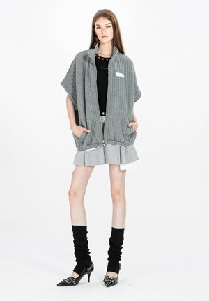 Joven con un suéter gris grande con cremallera, top negro, falda gris plisada, calentadores de piernas negros y tacones puntiagudos negros, de pie frente a un fondo blanco.