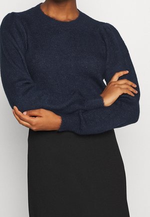 Osoba ubrana w ciemnogranatowy dziany sweter z bufkami na ramionach i długimi rękawami, w połączeniu z czarną spódnicą, z rękami skrzyżowanymi na wysokości pasa.