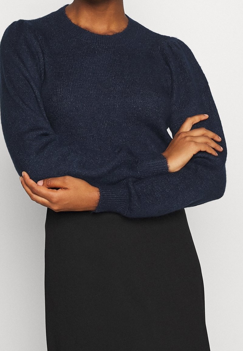 Personne portant un pull en tricot bleu marine foncé avec des épaules bouffantes et des manches longues, associé à une jupe noire, les bras croisés au niveau de la taille.