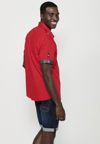 Camiseta roja de manga corta con detalles en denim y botones, combinada con shorts oscuros de denim con dobladillos enrollados y bolsillos laterales.