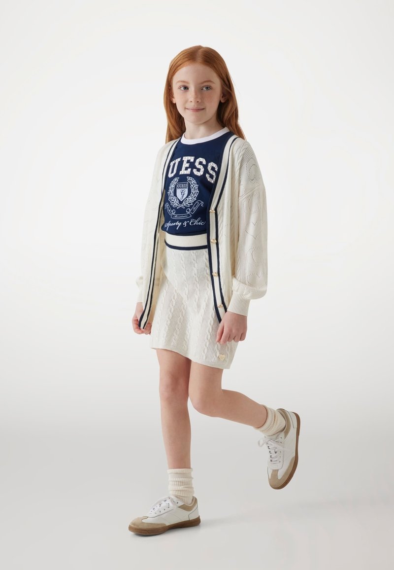Cardigan en tricot blanc sur un t-shirt graphique marine avec le logo "GUESS", associé à une jupe tricotée blanche et des baskets blanches avec des accents beiges.