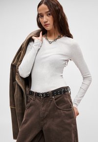 Šviesiai pilkas ilgomis rankovėmis marškinėlis, derinamas su rudos spalvos laisvo stiliaus kelnėmis ir juodu diržu su sidabriniais žiedeliais, viršuje dėvima ruda striukė.